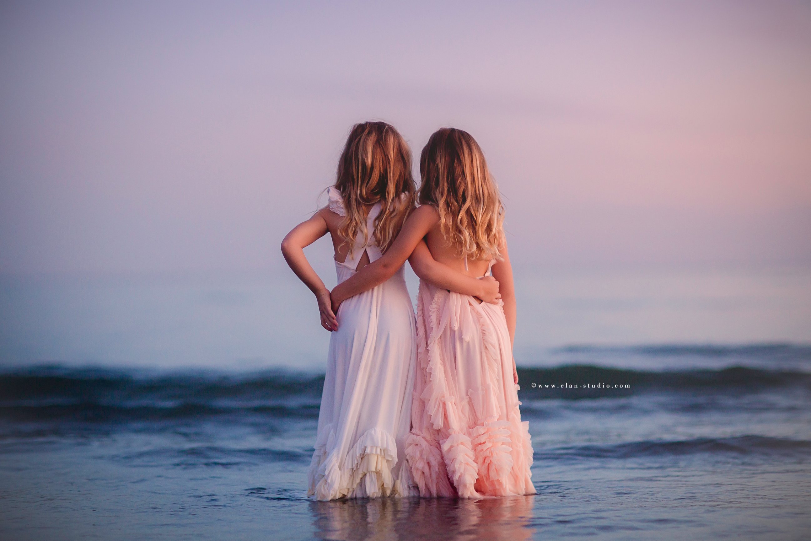 twin girls with arms around eachother, standing in ocean at sunset