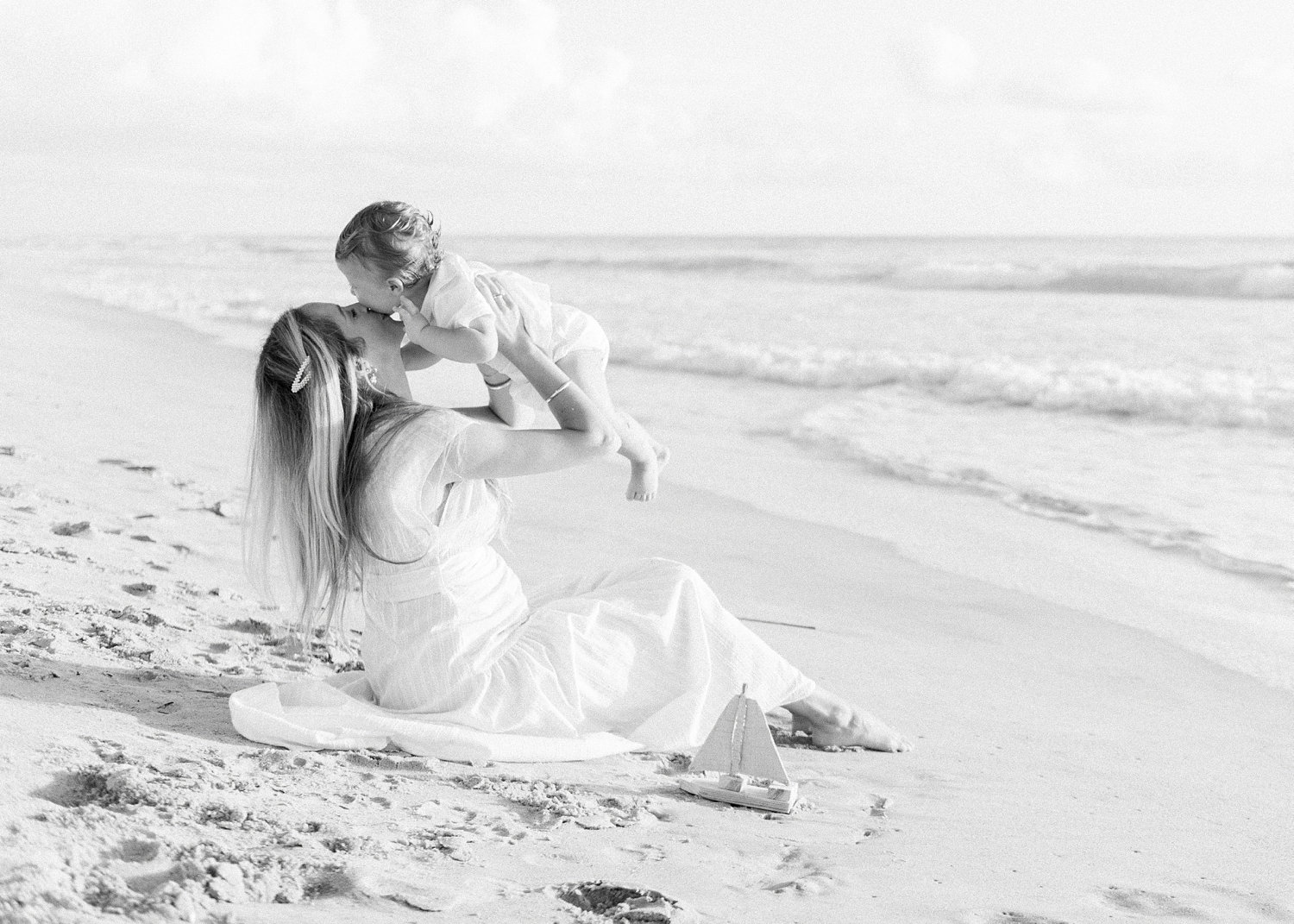 black and white photograph of a mother lifting up her baby boy to kiss him in the sand