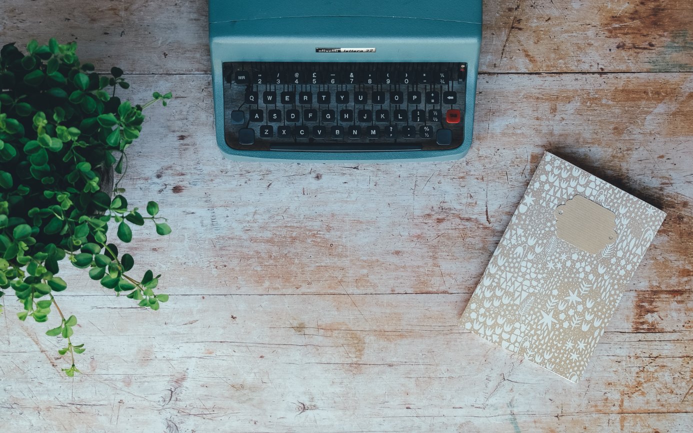 Vintage typewriter next to a patterned notebook and a small potted plant on a wooden tabletop.