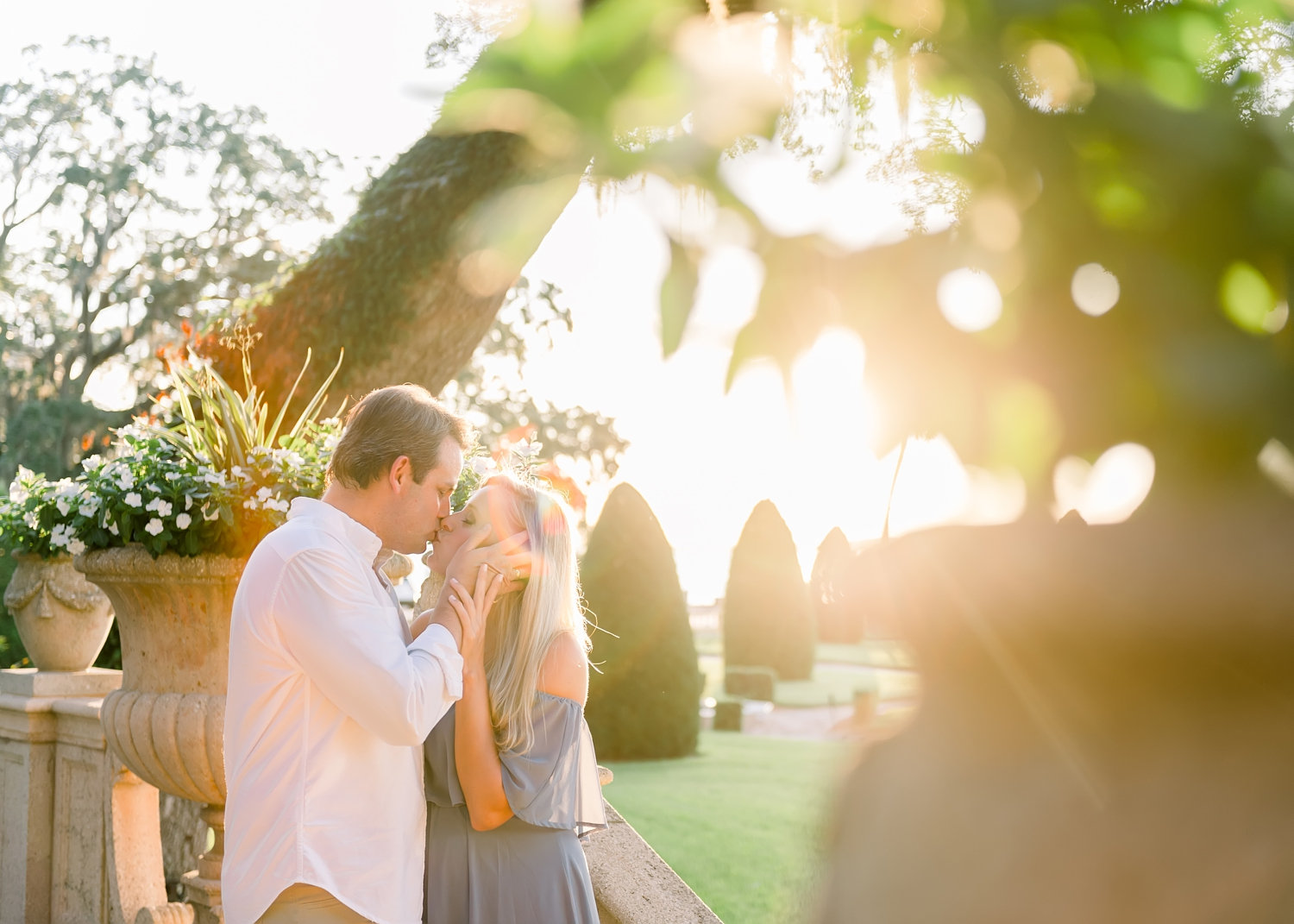 peeking through a large potted plant at a couple kissing with the sun setting behind them