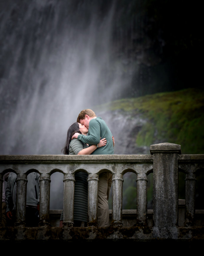 A couple embraces and kisses on a stone bridge with a waterfall in the misty background.