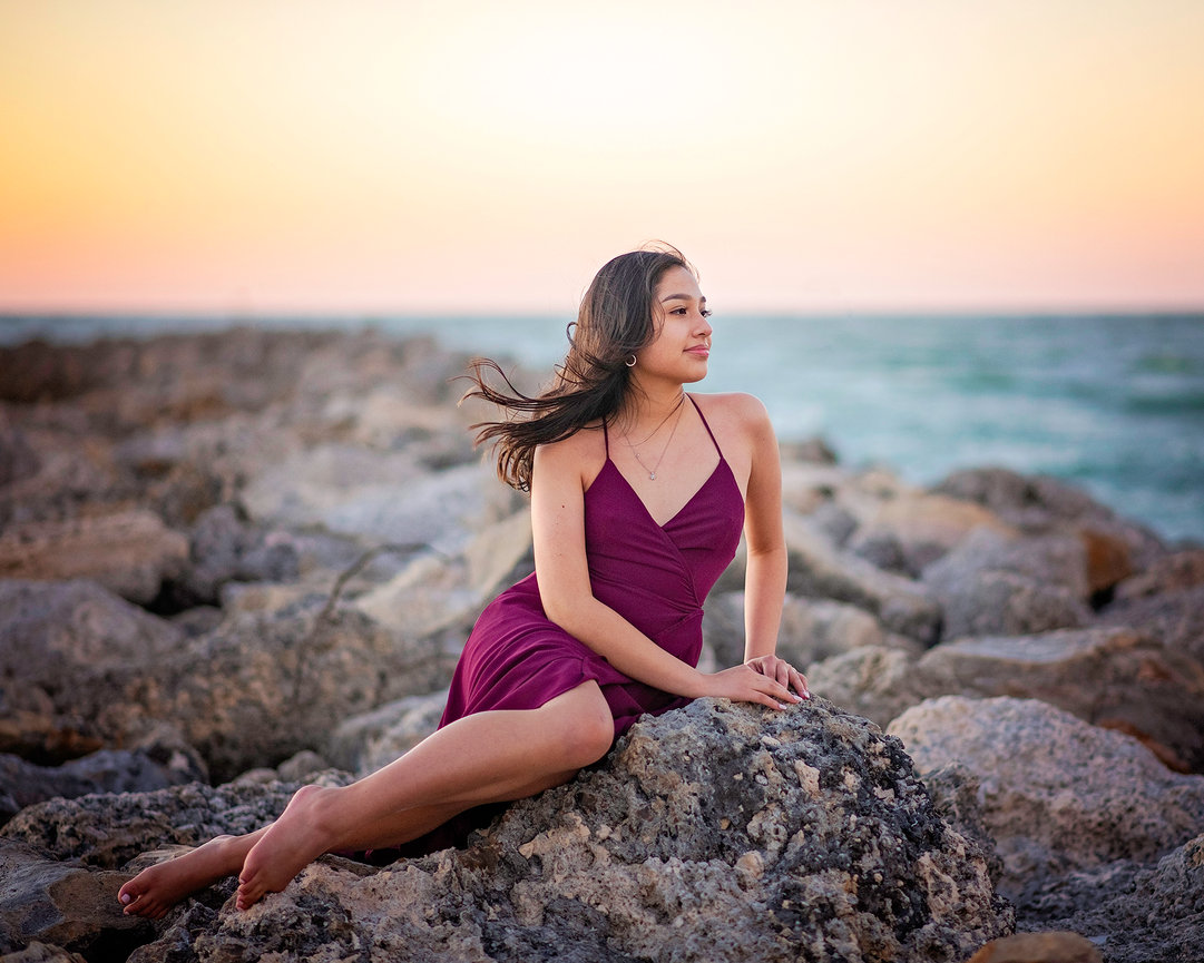 High school senior sitting on rocks with wind blowing hair on beach in St. Pete, FL.