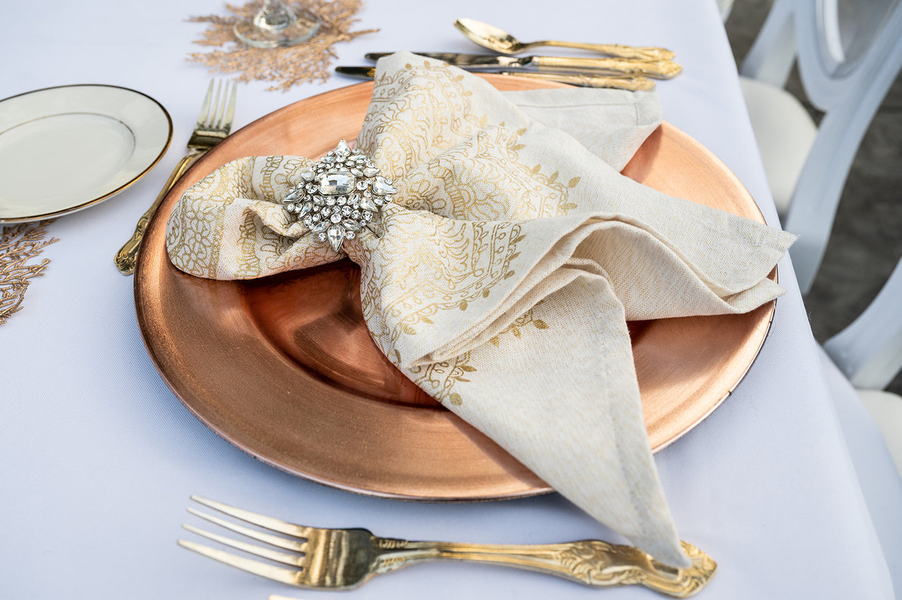 Elegant table setting featuring a copper plate, ornate napkin tied with a decorative brooch, and vintage silverware.