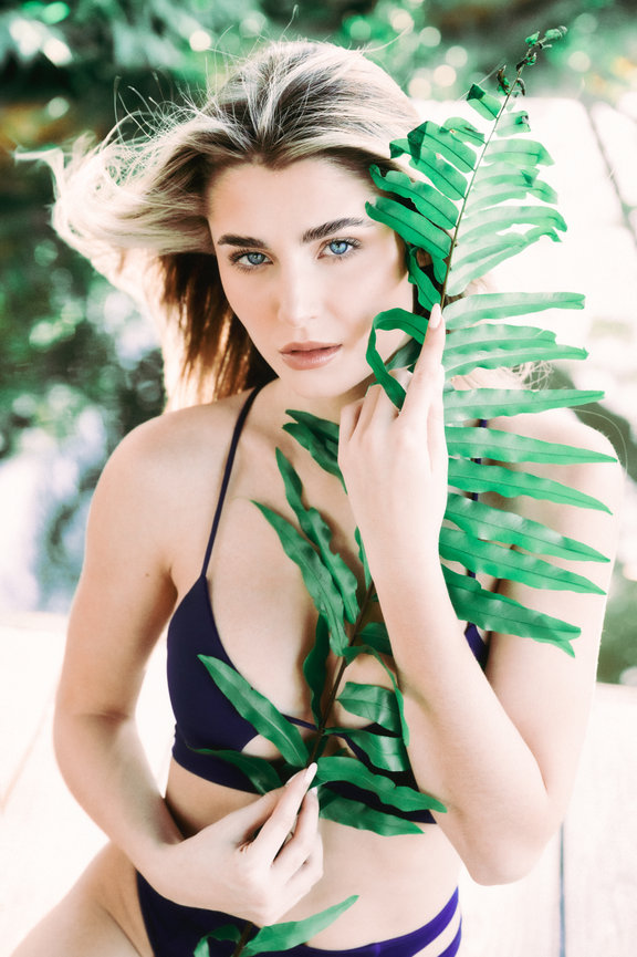 Model in a bikini poses with green leaves, showcasing a serene outdoor setting and striking blue eyes.