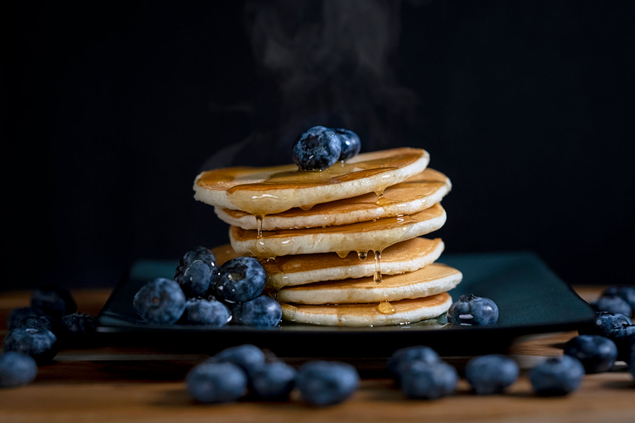 Stack of warm pancakes drizzled with syrup, garnished with fresh blueberries, against a dark background.