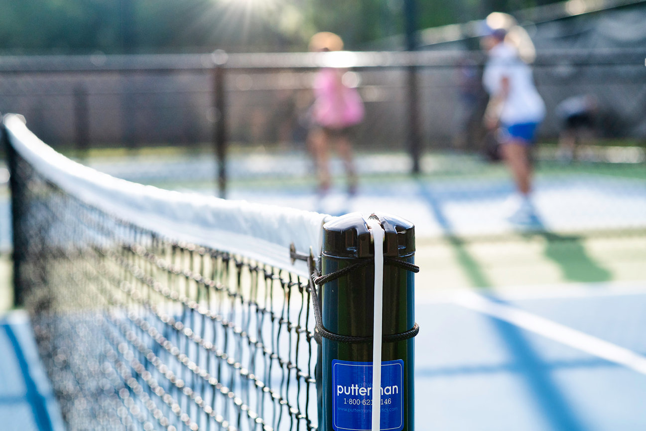 Close-up of a tennis net post with players in the background, enjoying a sunny day on the court.