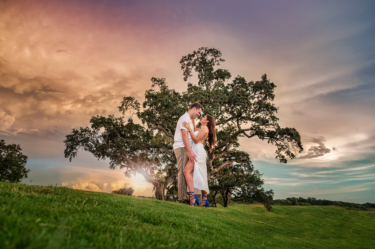 Couple embracing in a lush green field with a dramatic sunset in the background.