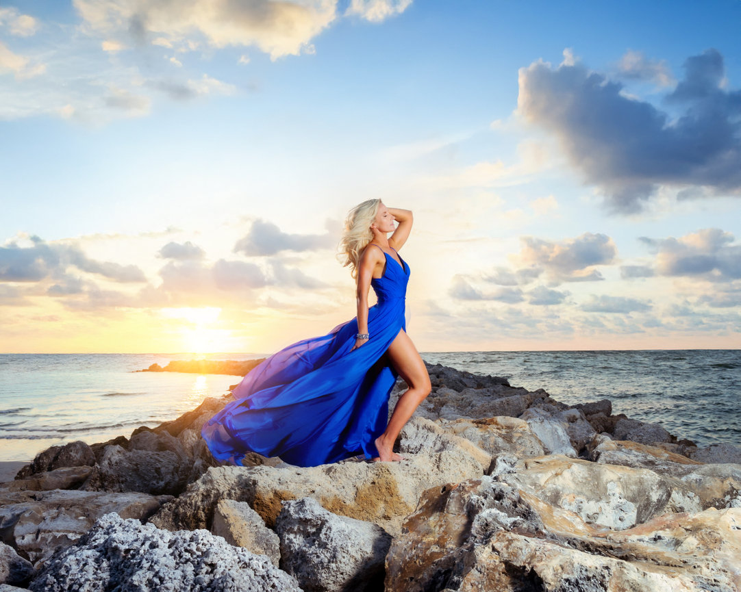 Woman in a flowing blue dress standing on rocks by the ocean at sunset, with clouds in the sky.