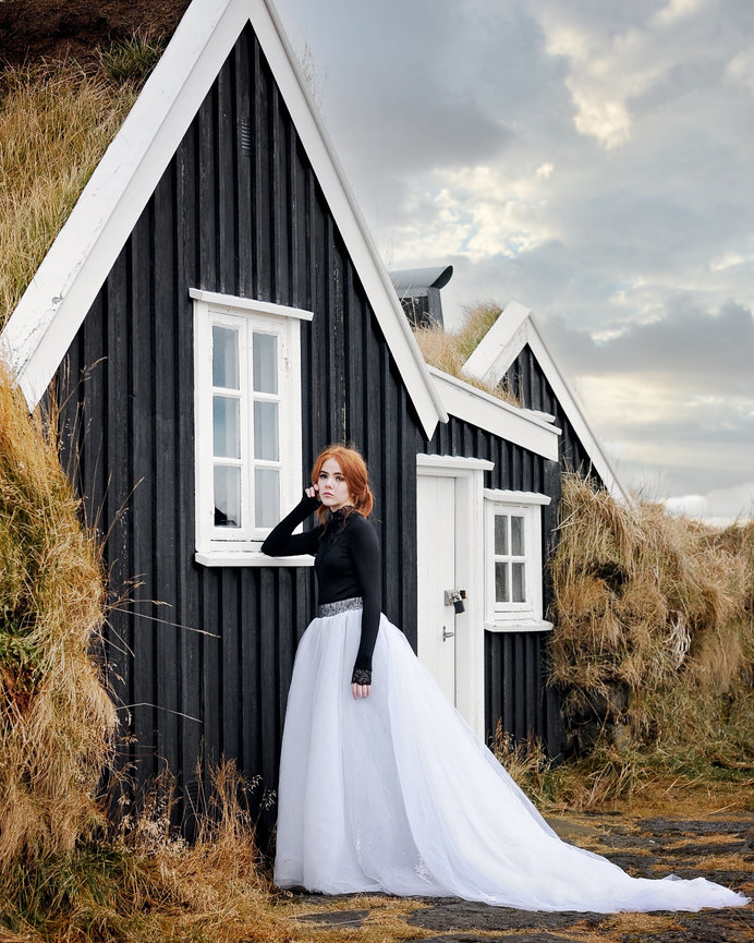 Woman in a flowing white gown poses near a black wooden house with grassy roof under a cloudy sky.
