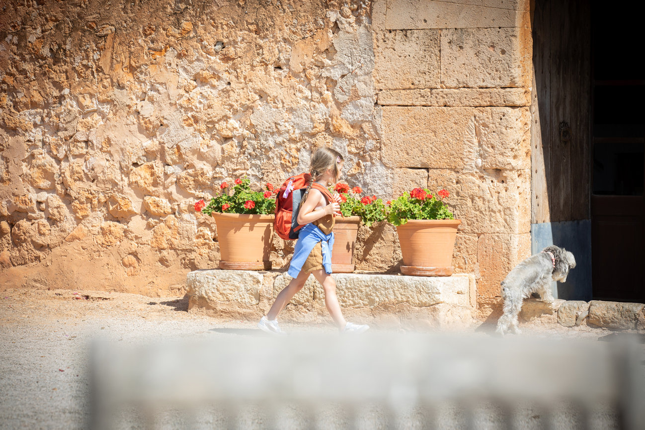 Child with backpack walking past potted flowers and a small dog near a rustic stone wall. Mallrca