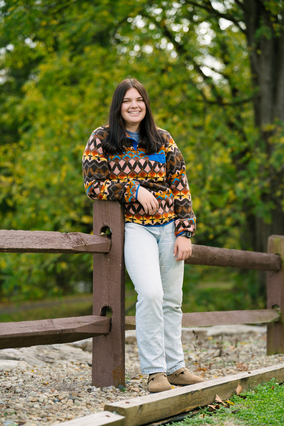 high school senior leaning against a fence in her senior pictures wearing a casual jeans and sweater outfit