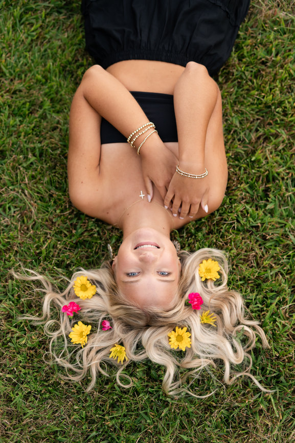 senior girl lying on grass, blonde hair adorned with yellow and pink flowers, wearing a black outfit and bracelets in clarkston mi park