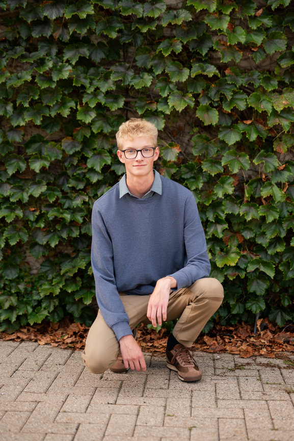 senior guy smiles softly in glasses kneeling on a brick path, wearing a blue sweater and khakis, with a leafy wall in the background in downtown clarkston mi