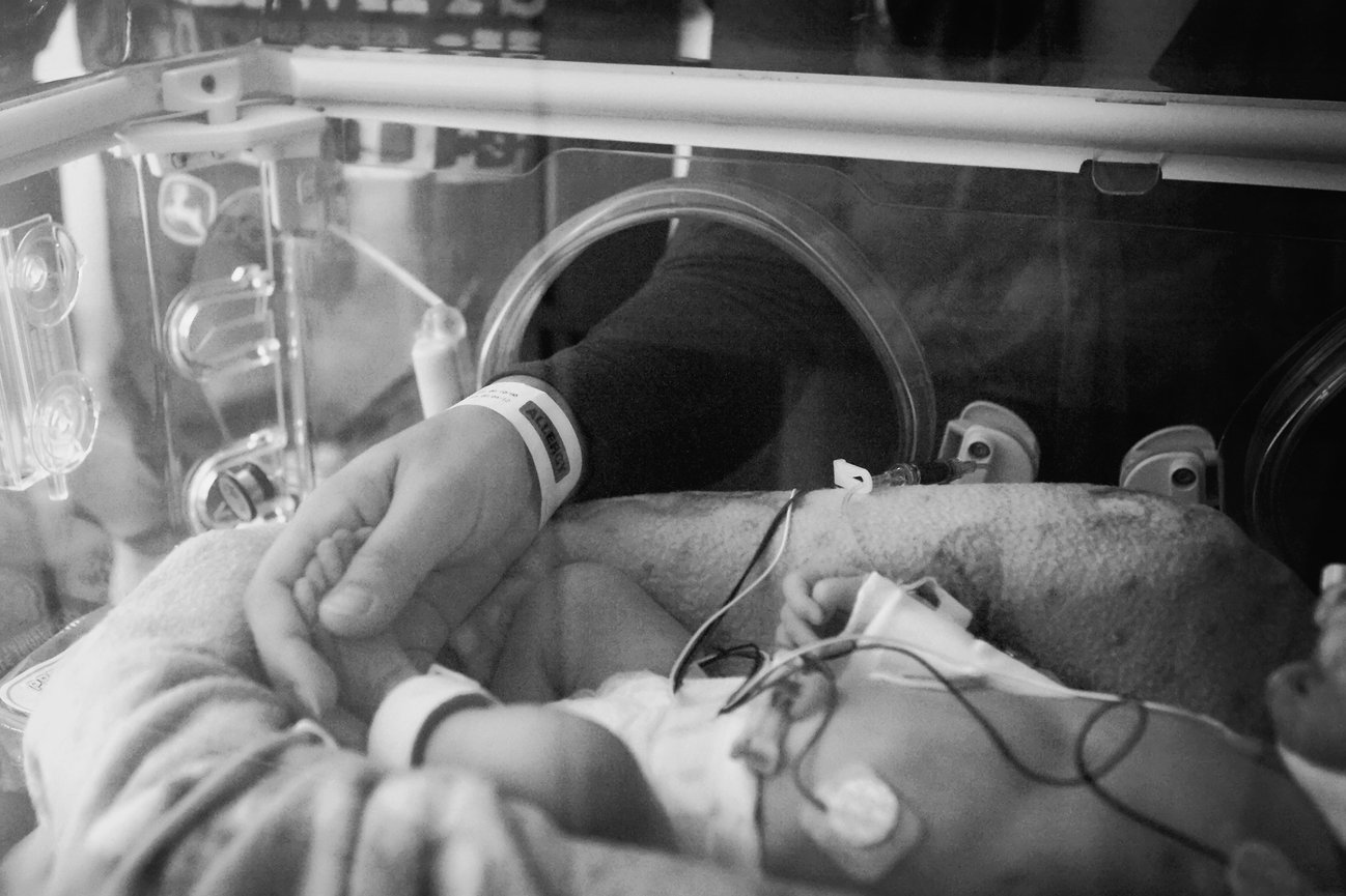 Adult holding hand of a baby in an incubator, both wearing hospital bracelets.