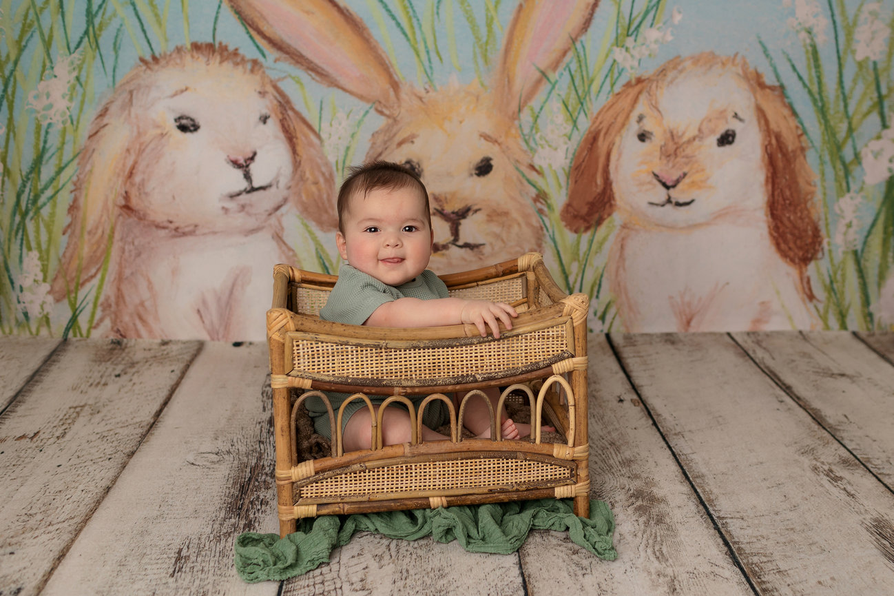 A baby smiling in a woven basket, with a backdrop of painted rabbits and grassy scenery.