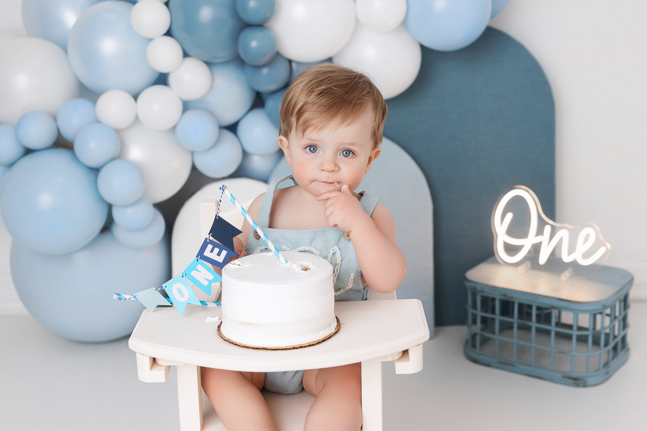 Baby in a high chair with a cake, surrounded by blue and white balloons, with a One sign lit up.