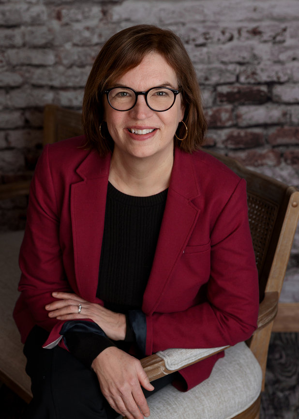 A person wearing glasses and a red jacket sits on a chair against a brick wall background, smiling at the camera.