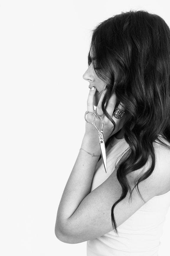 Woman with wavy hair holding scissors near her face, wearing a sleeveless top.