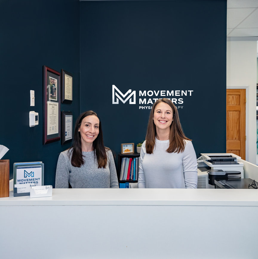 Two women smiling behind a reception desk at Movement Matters Physical Therapy.