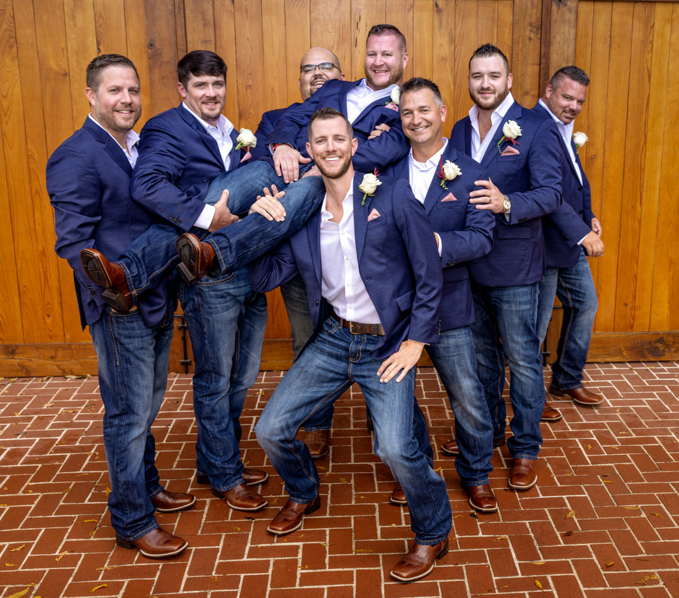 Groomsmen posing playfully in navy jackets and jeans against a wooden barn backdrop during a laid-back Alabama wedding.