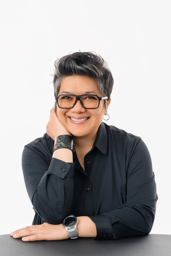 woman smiling, wearing glasses and a black shirt, seated with hand on chin, showing a tattooed wrist and watch.
