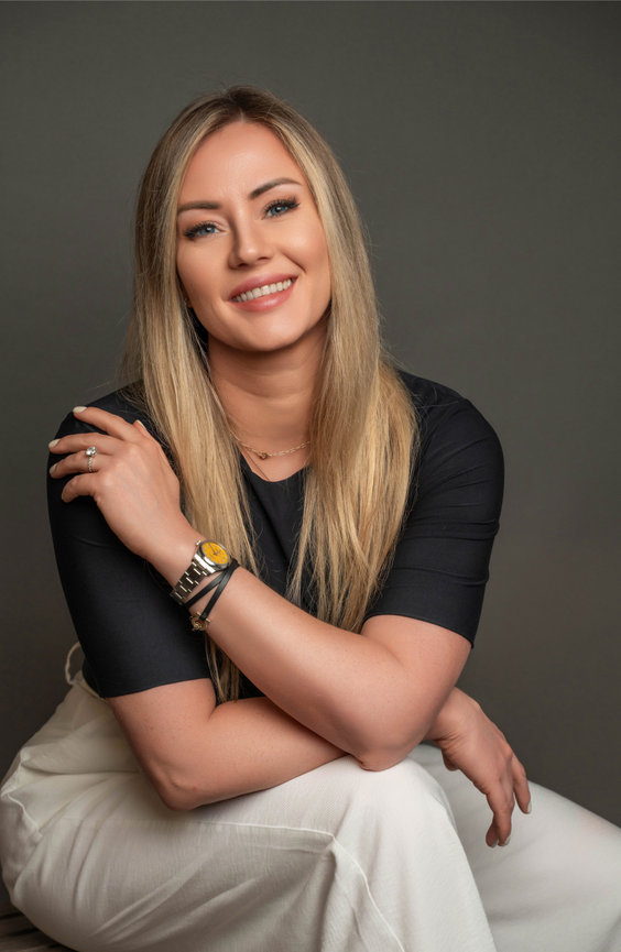 Woman seated in black top and white pants, smiling confidently.