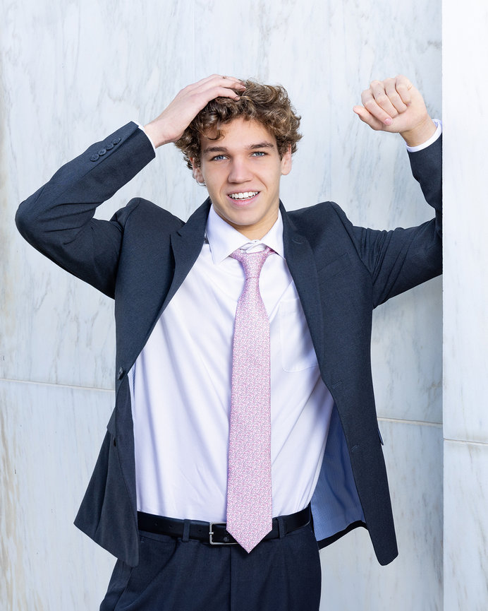 Guy in blue suit with white shirt and pink tie leaning on a white marble wall.