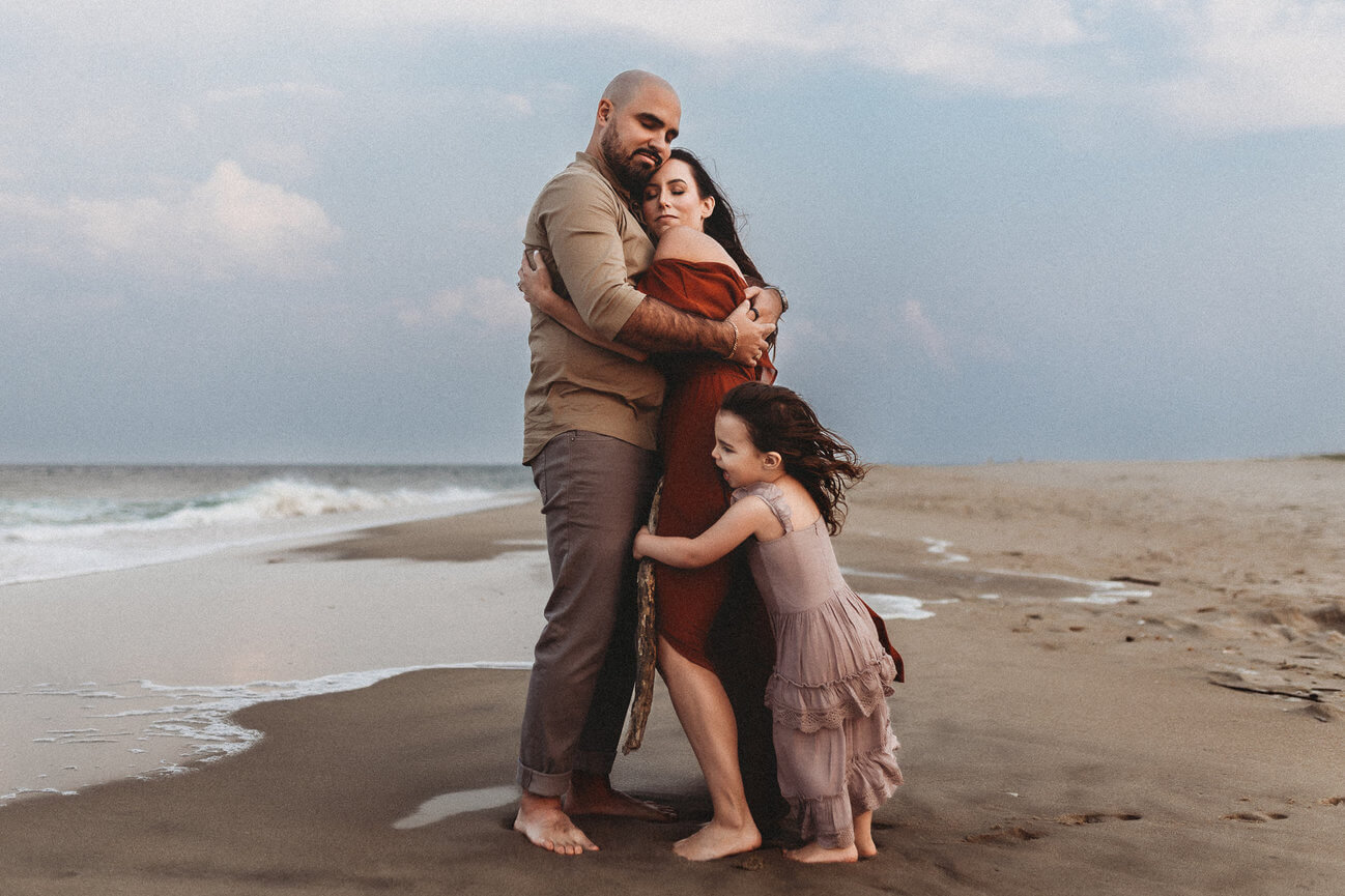 A family hugging tightly on the beach on a cloudy evening in Sandy Hook, New Jersey during their family photography session.