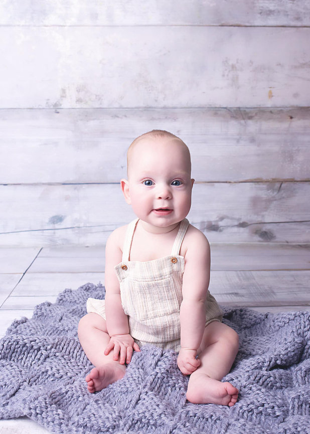 A blue eyed six month old baby sits on a grey knit blanket. The walls and floor are white wood.