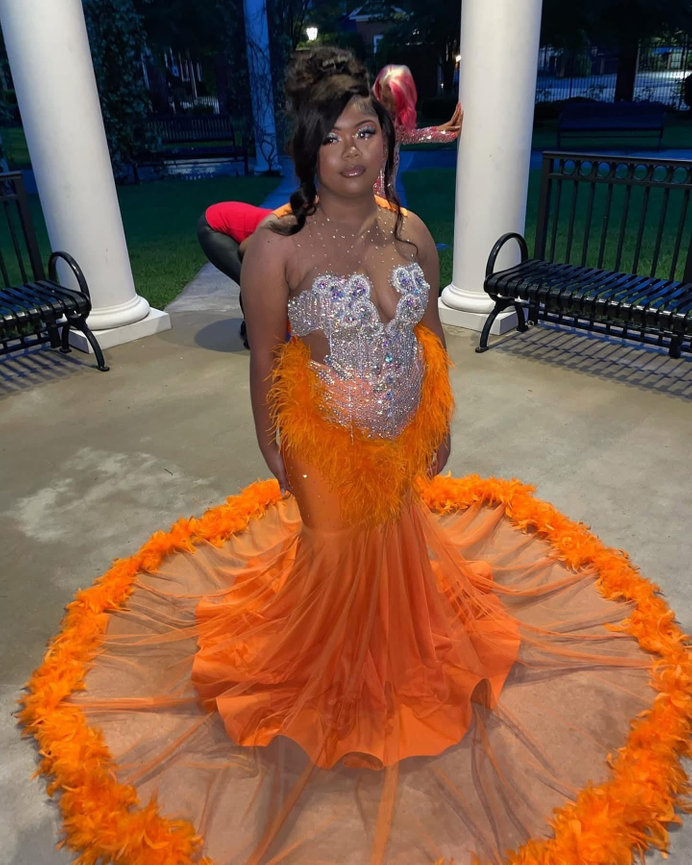 Person in an orange dress with feather accents stands on a porch, with a bench and greenery in the background.