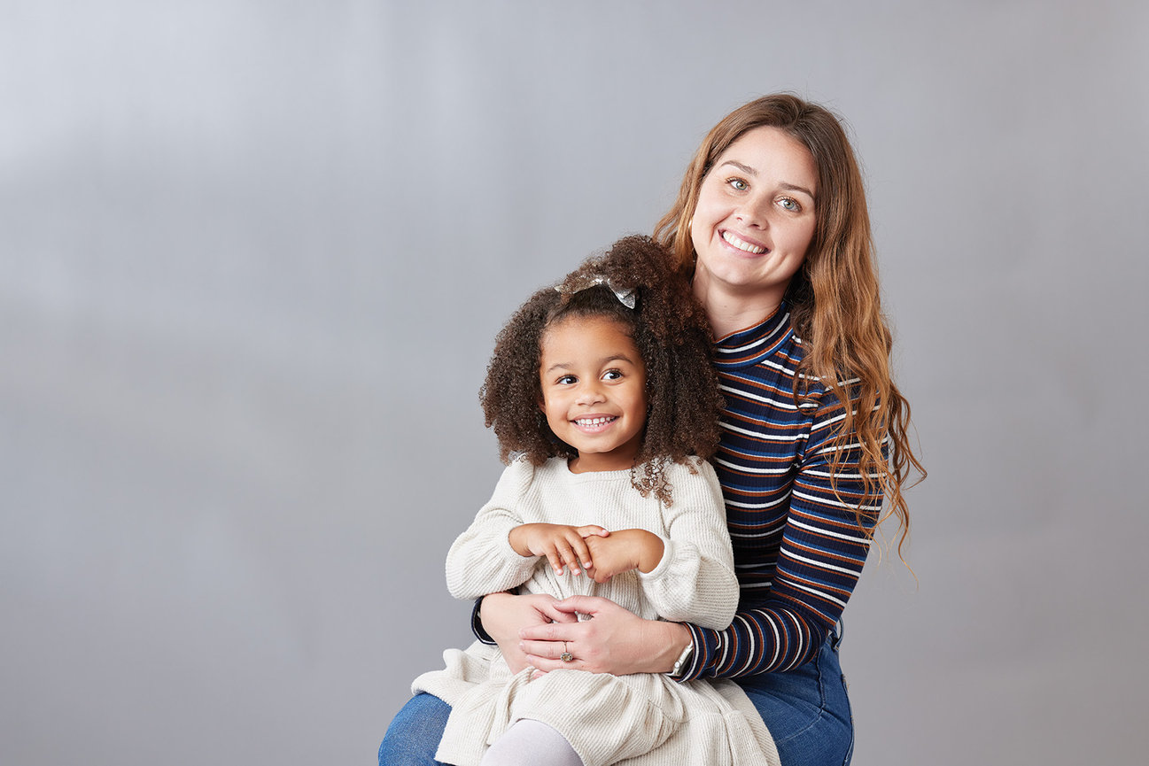 Indoor portrait of a mother and young daughter smiling together against a gray background in Carmel, Indiana, captured with soft natural-style light and a warm, connected feel