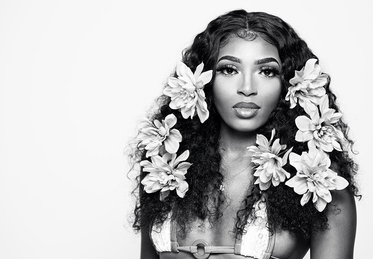 Black and white indoor portrait of a confident young woman with floral accents in her hair, soft lighting highlighting beauty, elegance, and a refined fine art look.