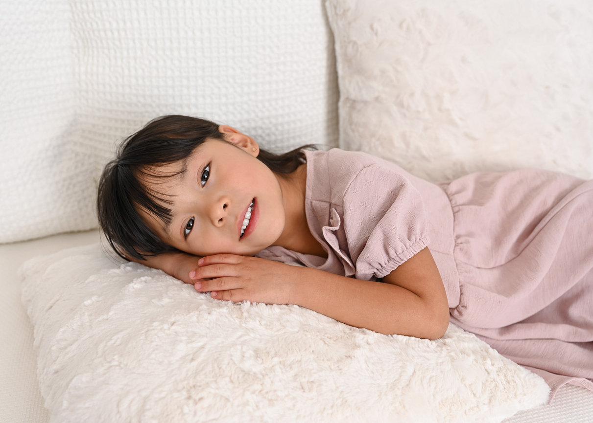 Child in a pink dress lying on white cushions, smiling at the camera.