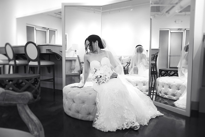 Bride in a strapless gown sits on a round ottoman in a dressing room with mirrors and a vanity.