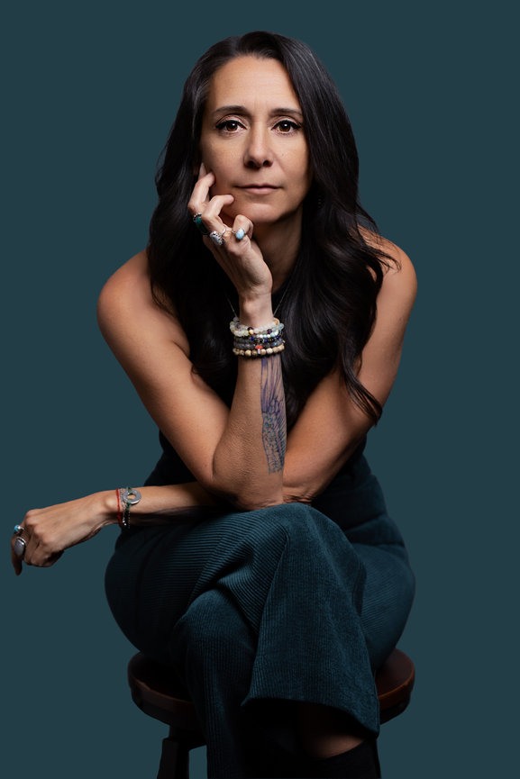 Woman with long dark hair seated, resting her chin on her hand, wearing bracelets against a dark teal background.
