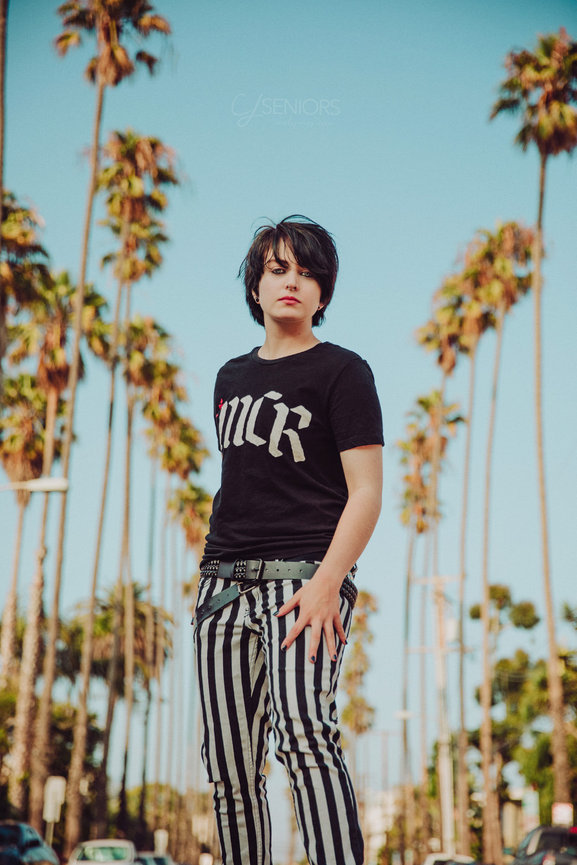 A goth teen boy standing in an iconic los angeles palm tree lined street in Santa Monica, CA, wearing band clothes by chele jones photographty