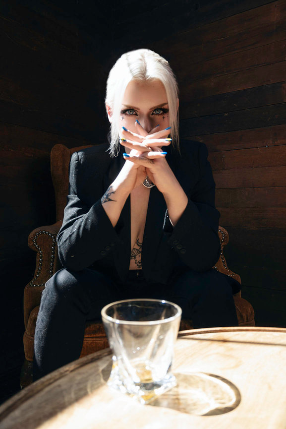 Empowered model seated in a Lehi, Utah studio, wearing a bold black suit with striking makeup and tattoos, exuding confidence and sophistication. A glass on a rustic table in the foreground, captured by Tre Nelson Photo.