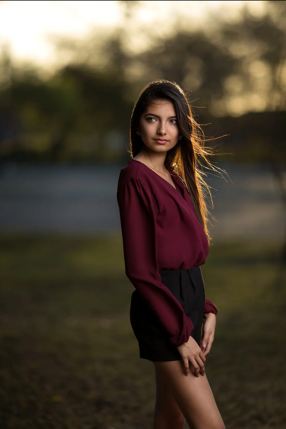 Woman in a burgundy blouse stands outdoors with a blurred background.