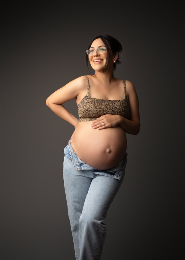 Pregnant woman in a leopard print top and jeans, smiling against a dark backdrop.