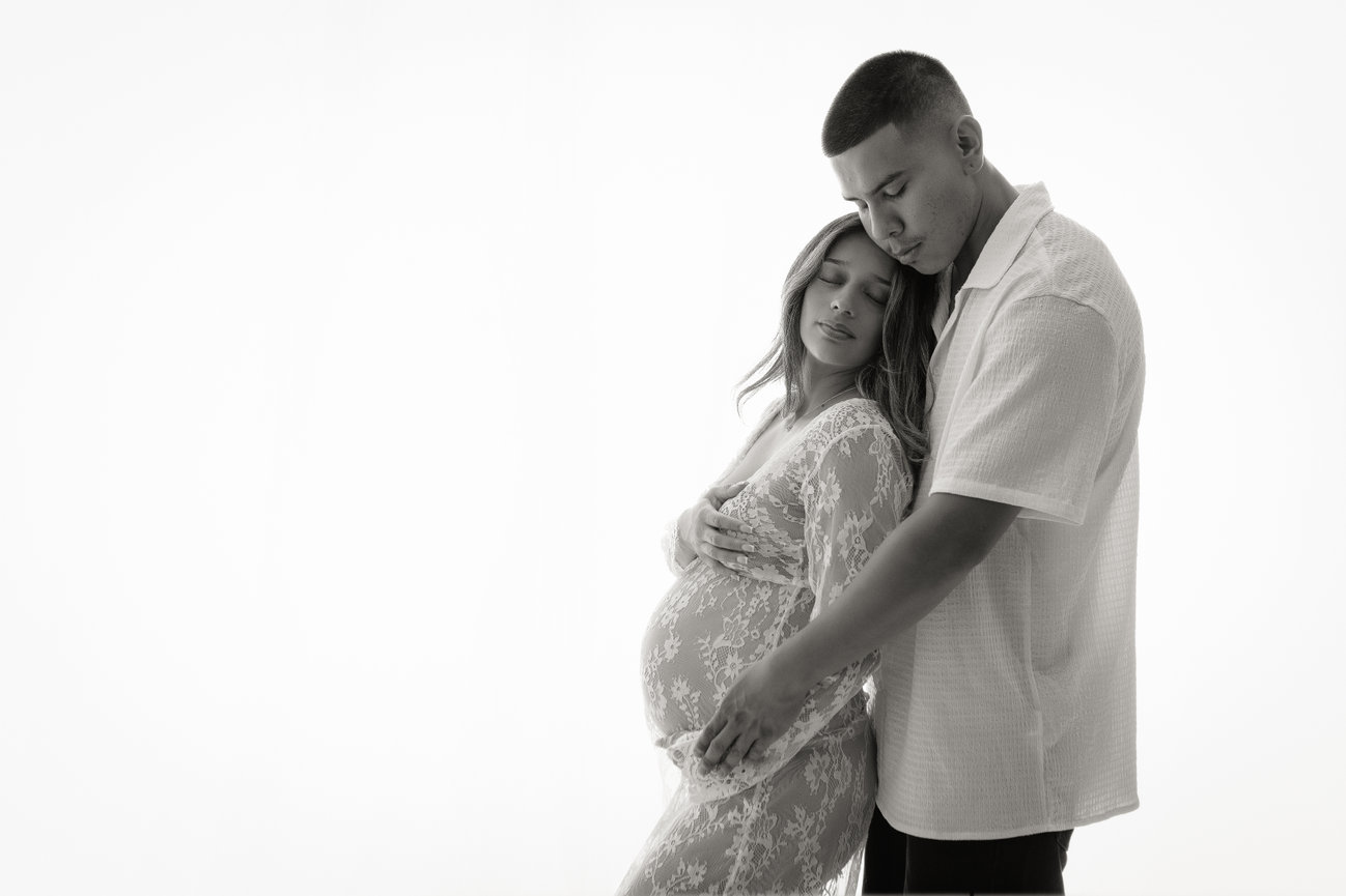 A pregnant couple in a loving embrace with the woman in a lace dress against a white background.