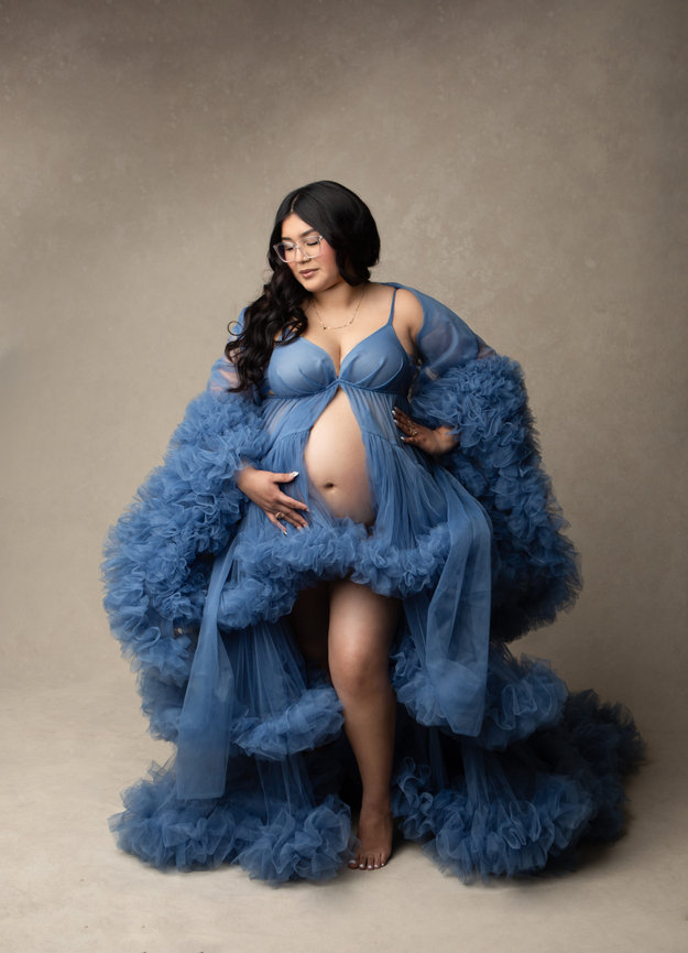 Pregnant woman in a flowy blue gown poses against a neutral backdrop, cradling her belly.