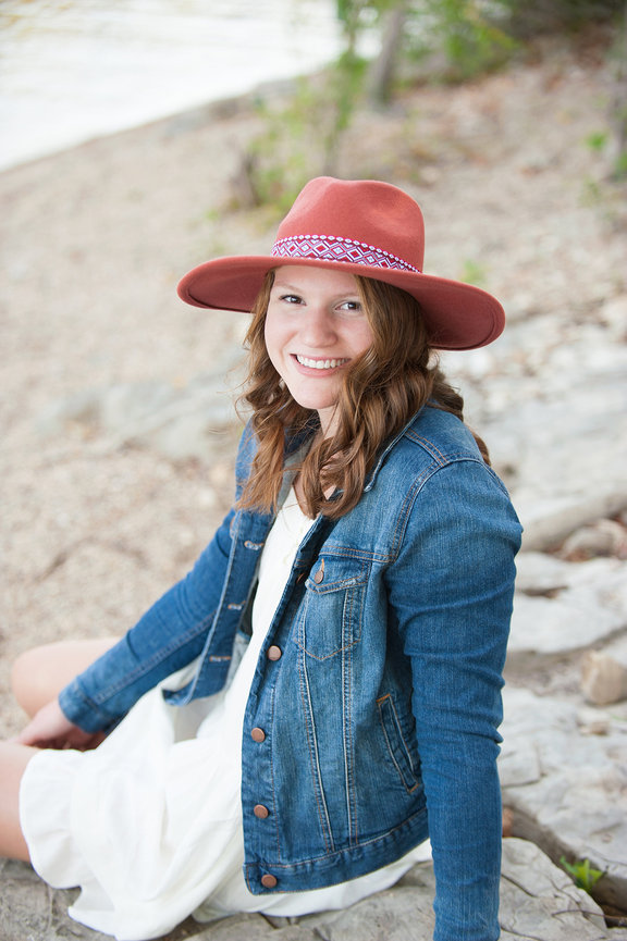 senior girl in jean jacket, white dress, and coral hat sitting on a rock at Table Rock Lake