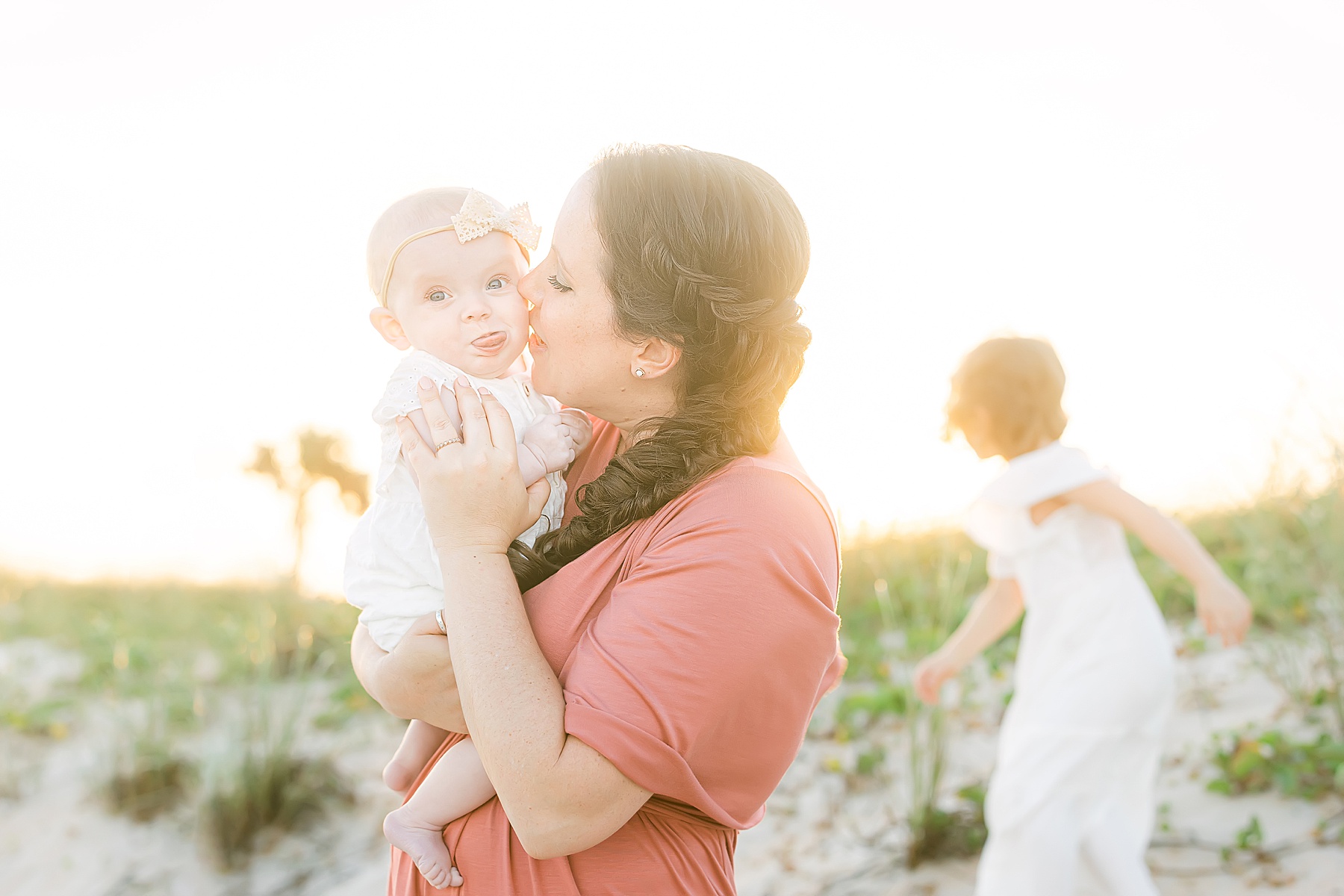 mom holding baby in white dress with braid in her hair on the beach