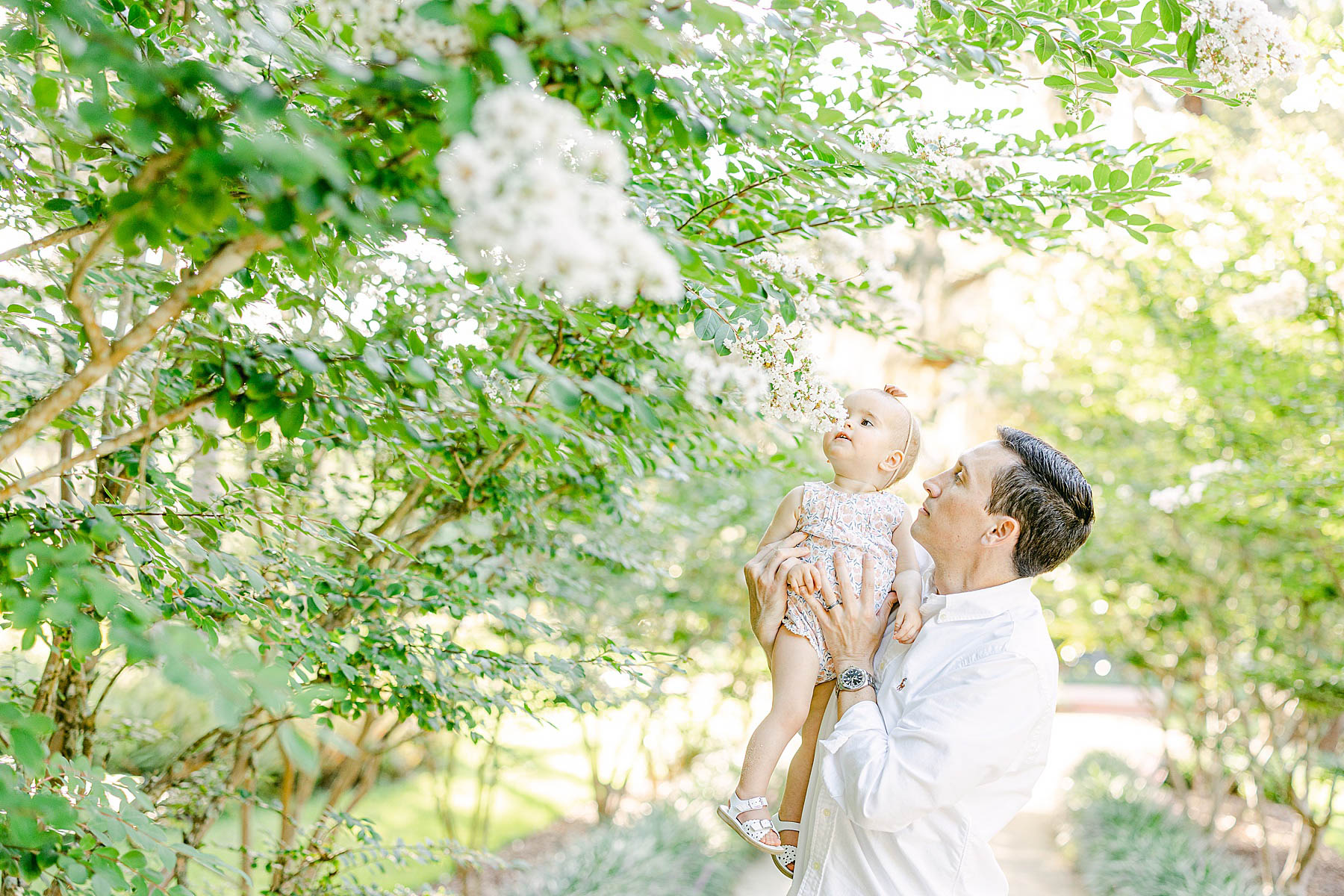 man holding baby girl in the air smelling white flowers