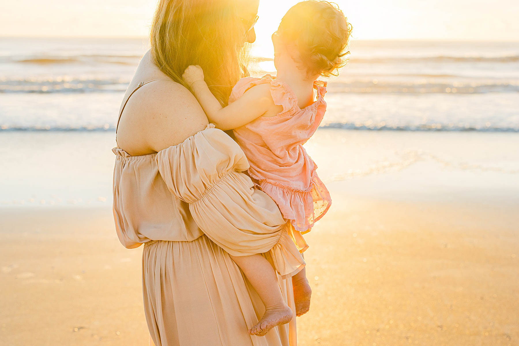 woman wearing taupe dress holding baby girl on the beach at sunrise with bright orange colors