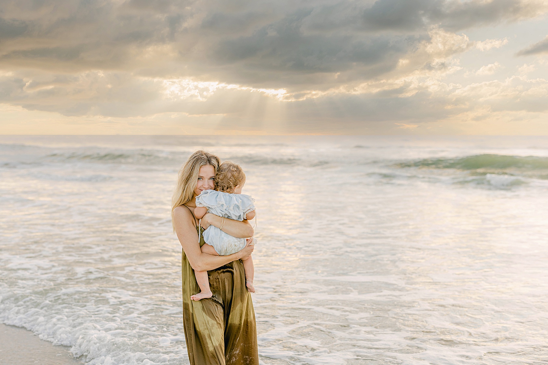woman with green eyes holding baby at sunrise on the beach standing in the water