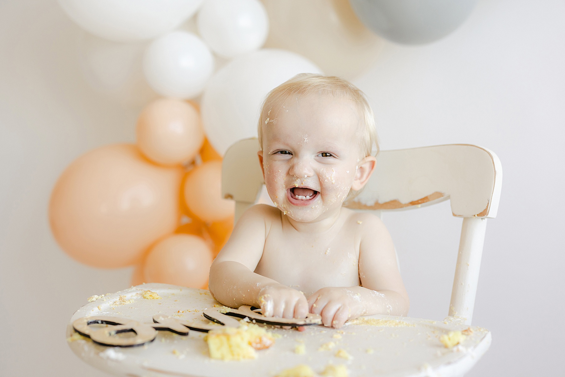 baby boy with cake all over his face sitting in white high chair with balloons in background