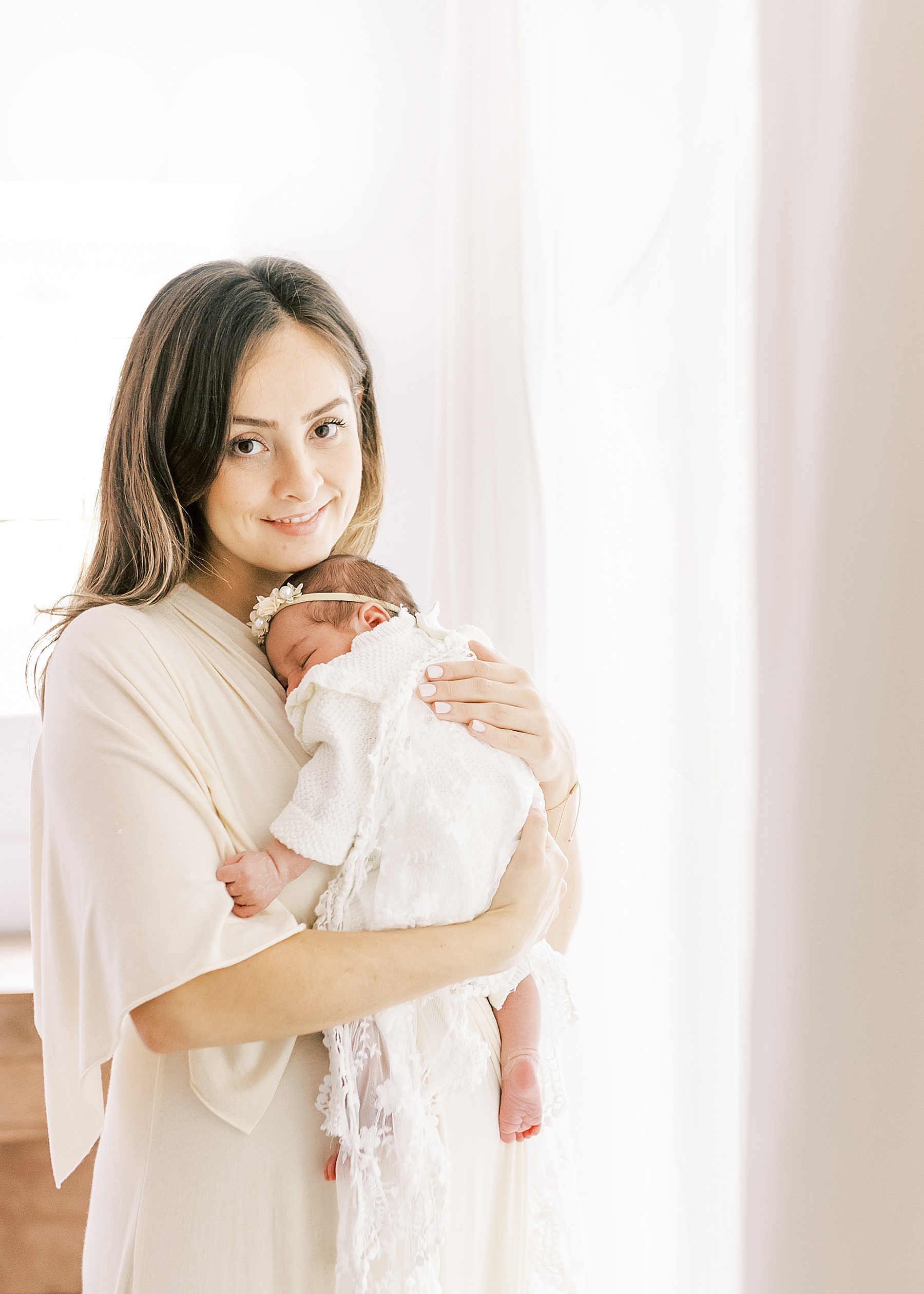 woman in cream long dress holding newborn baby girl dressed in white against backlit window white sheers