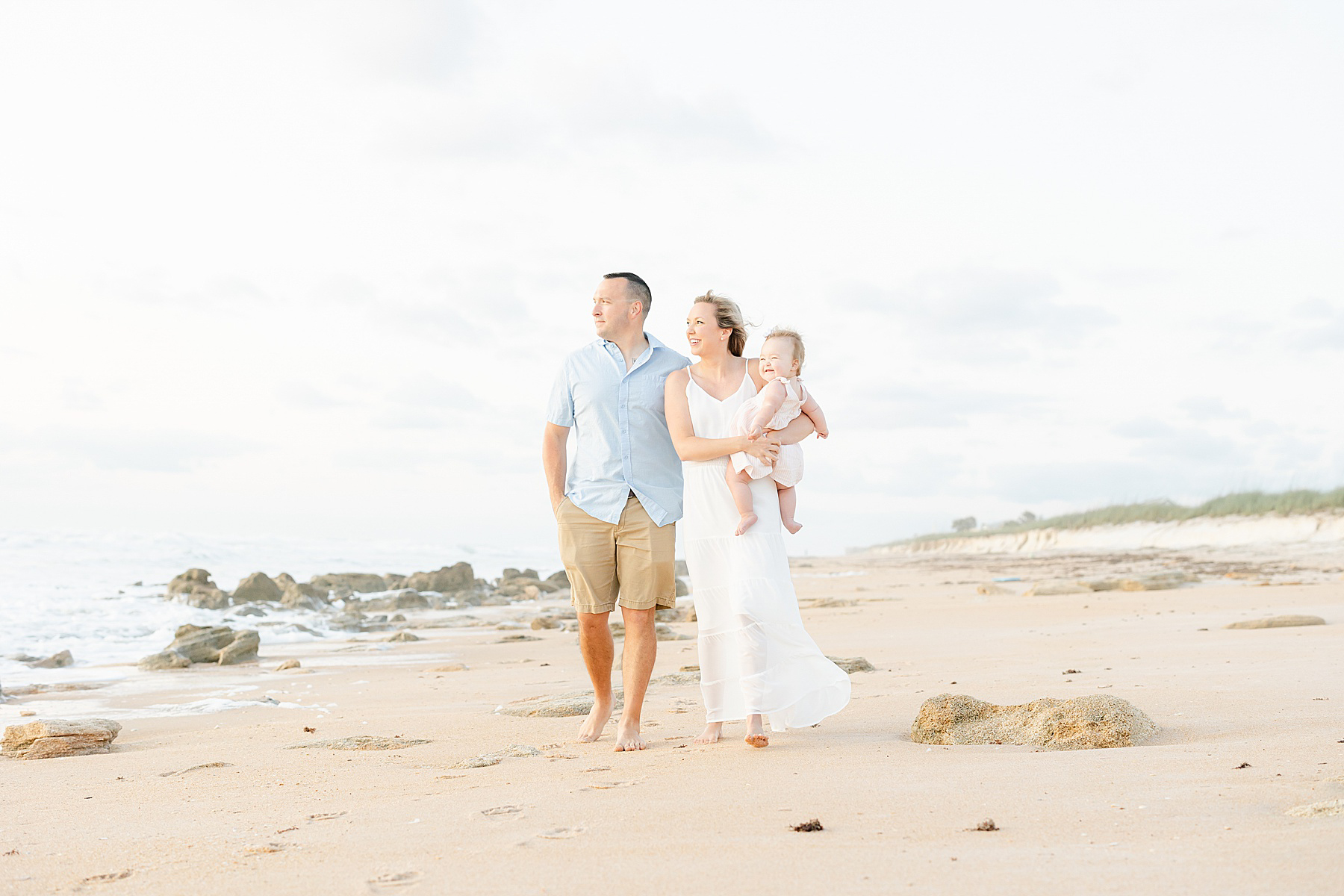 family of three walking on the beach at sunrise holding baby girl wearing pink striped dress