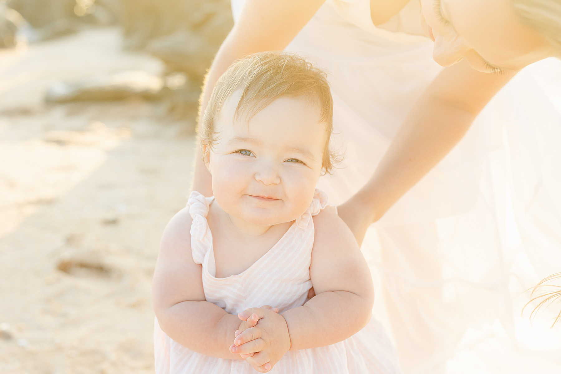 baby girl wearing pink and white striped dress holding her mama on the beach